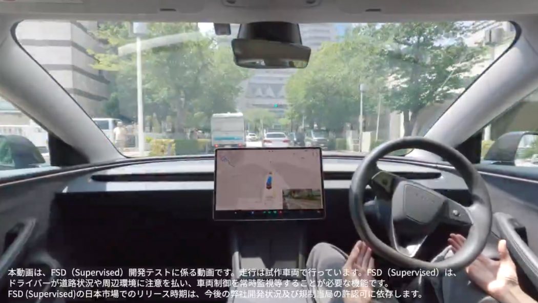 テスラFSD自動運転テスト走行の車内映像
テスラ車の運転席から撮影された映像で、ドライバーの両手がハンドルから離されており、自動運転中であることを示しています。右側にはテスラ特有の大型タッチスクリーンディスプレイが設置されており、画面には車両周辺の状況を示すグラフィカルな表示が映っています。
フロントガラス越しには日本の都市部の街並みが見え、街路樹が並ぶ道路、他の車両、建物などが確認できます。車内のインテリアはテスラらしいミニマルなデザインで、黒を基調とした内装となっています。
画面下部には日本語で以下のテロップが表示されています：
「本動画は、FSD（Supervised）開発テストに係る動画です。走行は試作車両で行っています。FSD（Supervised）は、ドライバーが適切な注意力を払い、周辺を常時監視し、必要な場合にはいつでも運転を引き継ぐ必要があります。FSD（Supervised）の日本市場でのリリース時期は、今後の許可申請状況及び規制当局の許可可に依存します。」
この映像は、テスラが日本でのFSD技術の実証実験を行っていることを示すものです。
