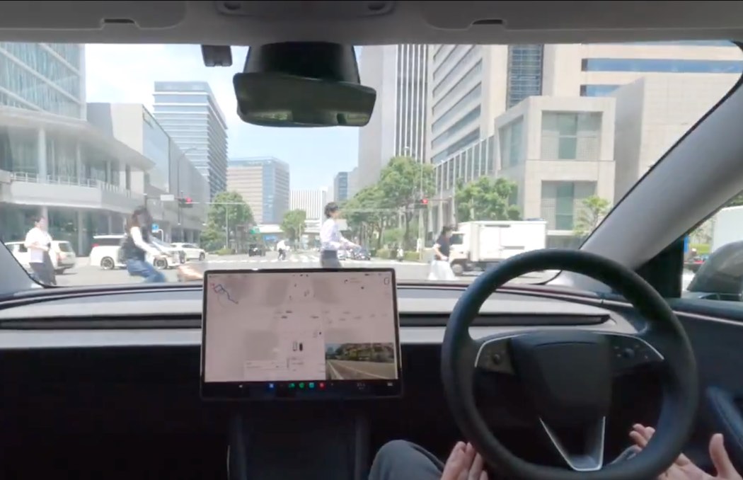 この画像は、テスラの運転席から撮影された写真です。ダッシュボード中央に大型のタッチスクリーンディスプレイが設置されており、地図アプリケーションが表示されています。運転者の両手がステアリングホイールに軽く添えられている様子が見え、フロントガラス越しには現代的な高層ビルやオフィスビルが立ち並ぶ都市部の街並みが広がっています。道路上には歩行者や他の車両が確認でき、晴天の日中に撮影された自動運転テストの様子です。