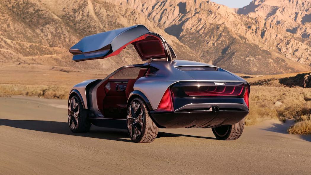 キャデラック・エレベーテッド・ベロシティ コンセプトカーの後方斜めからの撮影写真。ガルウィングドアが開かれ、赤い内装が見える状態のシルバーグレーの電動クロスオーバーSUV。特徴的な赤いLEDテールライト、24インチの大径ホイール、筋肉質なリアデザインが印象的。砂漠の未舗装路に駐車され、背景には乾燥した山岳地帯の雄大な景色が広がる。
