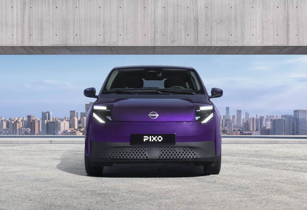 Photograph showcasing a purple Nissan Pixo concept car parked before a concrete architectural opening with a city skyline beyond. Front view highlights illuminated angular headlights, closed grille, PIXO plate, sleek compact design, and blue sky backdrop.